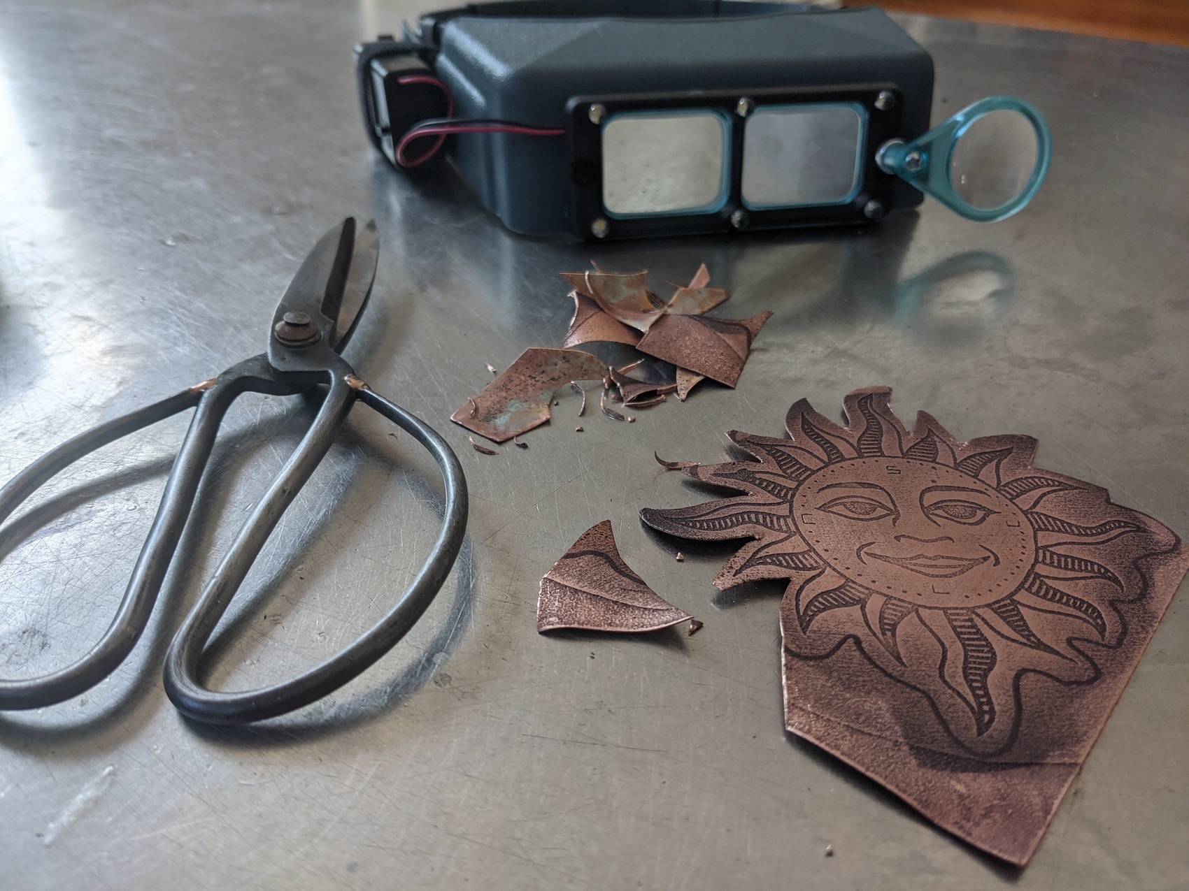 a piece of plate copper with s sun etched onto it, and bunch of scraps, and a sturdy pair of scissors lie on a stainless steel table